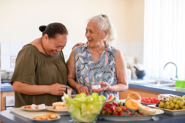 An elderly woman laughs alongside her carer as they prepare food.