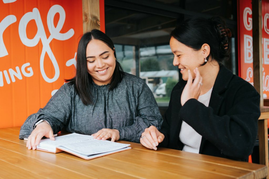 Two smiling women look at a notebook together.